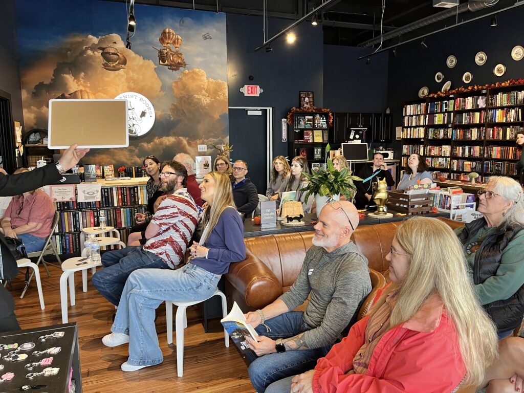 Seated crowd among the shelves watches Nick Gasparro hold up a reveal board during a Lockport mentalism show
