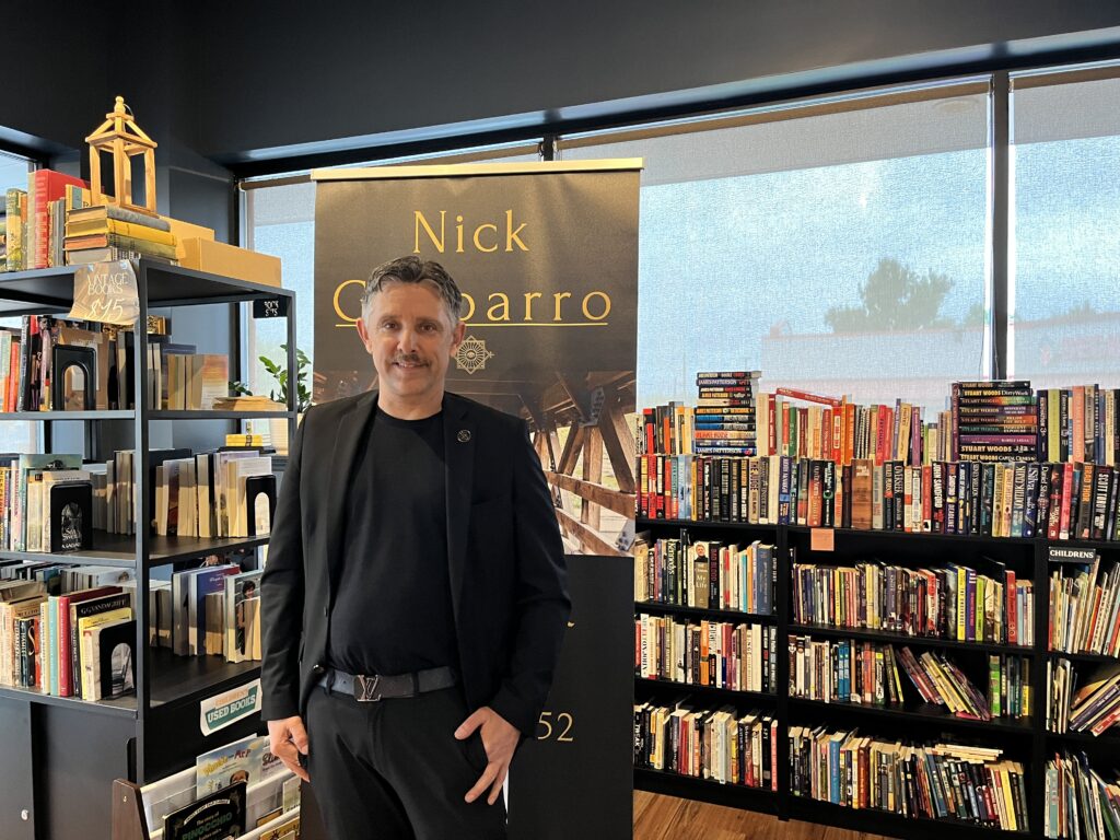 Magician and mentalist Nick Gasparro smiling beside his show banner among the bookshelves
