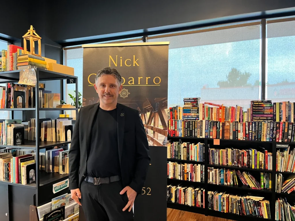 Magician and mentalist Nick Gasparro smiling beside his show banner among the bookshelves